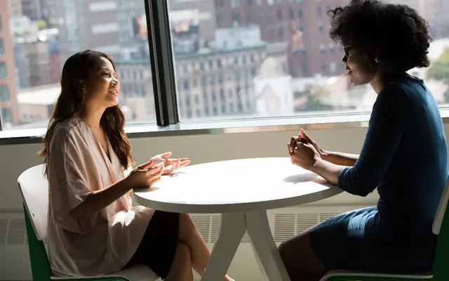 Dos personas conversando en una mesa junto a una ventana
